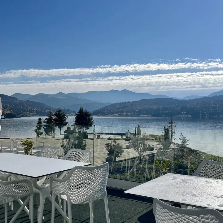 Mese și scaune albe pe terasa cu balustradă de sticlă, spre lac și munți